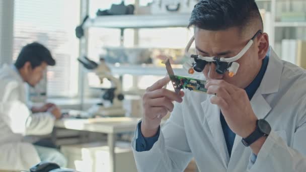 与男性同事一起在科学实验室工作的亚洲男性工程师身着白色实验室外套 通过镜片百叶窗看电路板 他的胸部动作缓慢 — 图库视频影像