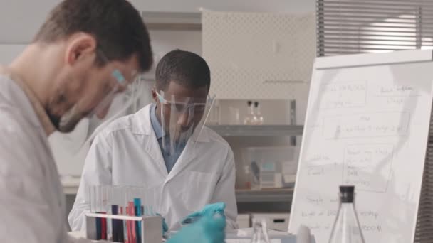 顔の盾と白い研究室のコートの2人の若い多民族男性化学者のウエストアップの低速物質に関する研究を行い 実験室での試験管内の異なるサンプルを研究 — ストック動画