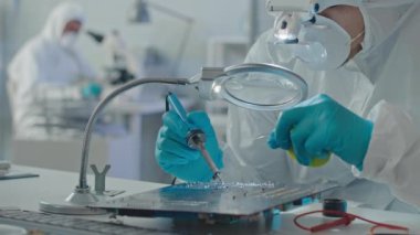 Beyaz koruyucu tulumlar, solunum maskesi ve lehim baskılı devre kartı takan erkek mühendisin belini kaldır ve büyüteçle ona bak.