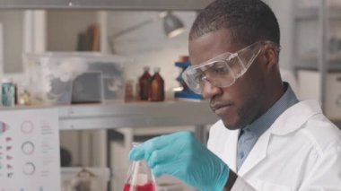 Koruyucu gözlük takan ve laboratuvar önlüğü giyen genç Afrikalı Amerikalı kimyagerin göğsü yavaşlıyor. Cam şişeyi kırmızı sıvı maddeyle karıştırıyor ve laboratuvarda araştırma yapıyor.