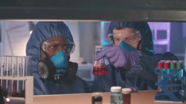 Koruyucu mavi tulumlu, solunum maskeli ve gözlüklü iki erkek biyoteknoloji uzmanının göğüs röntgeni. Cam test tüpleri ve gizli laboratuvar şişelerinde incelenen maddeler.