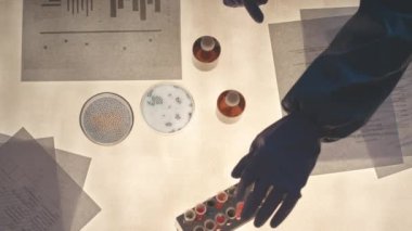 Üst görünüm, üzerinde doküman yığınları, test tüpleri ve petri kapları olan maddelerle araştırma yapan, koruyucu giysiler içinde tanınmayan iki biyoteknoloji uzmanının yavaşlaması.