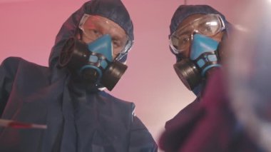 Koruyucu tulumlu, gözlüklü ve solunum maskeli iki erkek biyoteknoloji uzmanının, petri kabındaki maddeleri tartışırken ve incelerken karanlık gizli laboratuvarda araştırma yaparken düşük açılı yavaşlaması.