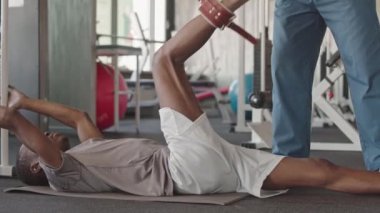 Modern tıp merkezindeki seans sırasında genç siyahi bir adamın kinezi simülatörü üzerinde rehabilitasyon bacak egzersizleri yapmasına yardımcı olan kişisel antrenör.