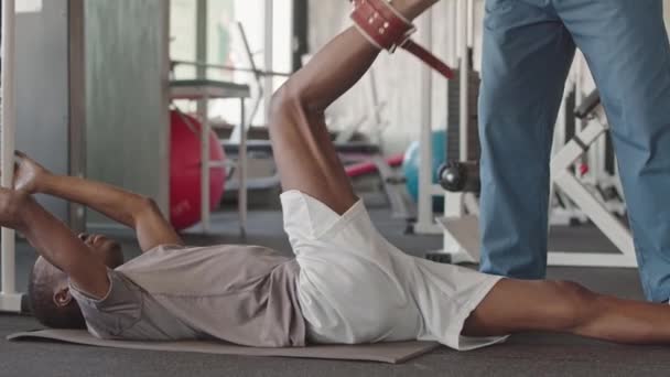 現代医療センターでのセッション中に運動療法シミュレータ上のリハビリテーションの脚の演習を行うために若い黒人男性を助けるパーソナルトレーナー — ストック動画