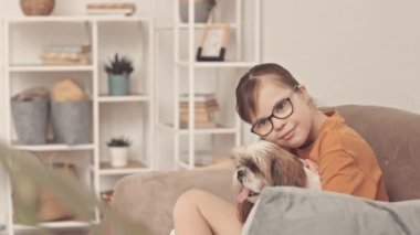 Oturma odasında kanepede oturan sevimli shih tzu köpeğini okşarken kameraya gülümseyen Down sendromlu küçük bir kızın portresi.