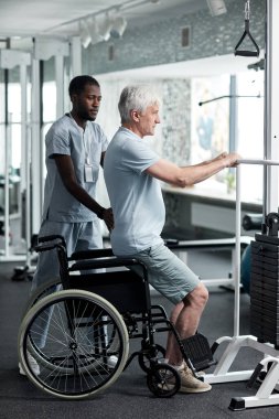 Tıbbi klinikte rehabilitasyon egzersizleri yapan yaşlı bir adamın tam boy portresi. Terapistin de yardımıyla.