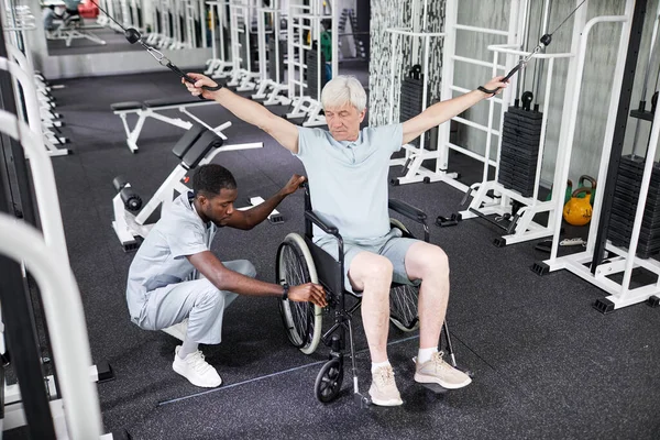 stock image Portrait of African American rehabilitation therapist assisting senior man in wheelchair doing physical exercises in gym