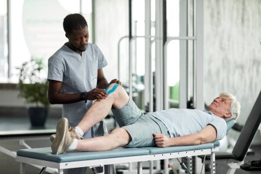 Rehabilitasyon terapistinin portresi fizyoterapi kliniğinde kıdemli bir hastanın dizine bant yapıştırıyor.