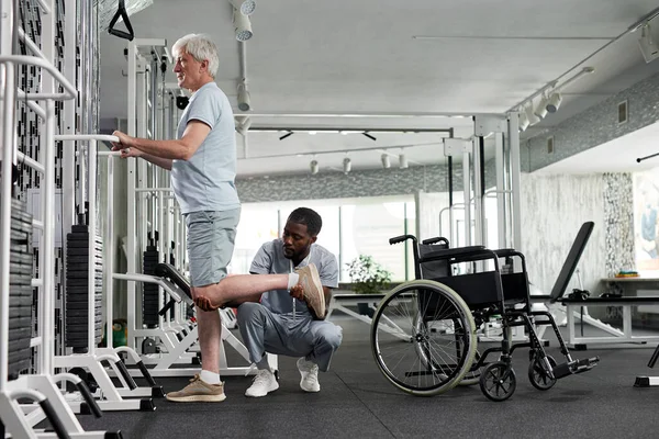 stock image Side view of senior man doing rehabilitation exercises in gym with therapist assisting, copy space