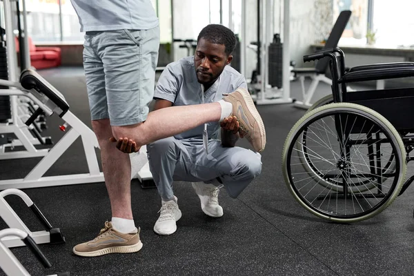 Portrait of African American rehabilitation therapist helping patient doing mobility exercises in gym, copy space