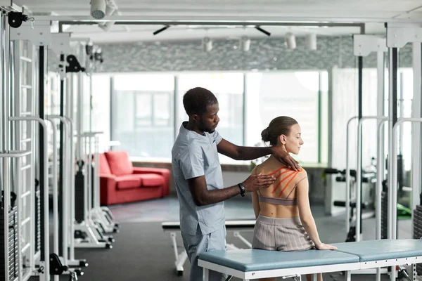 Rehabilitation therapist putting kinesio tape on back and shoulder of young woman in physiotherapy clinic, copy space