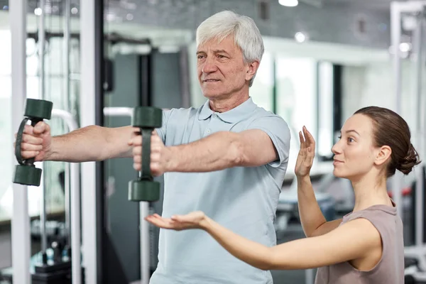 stock image Portrait of smiling senior man doing exercises in gym at rehabilitation clinic with female therapist assisting