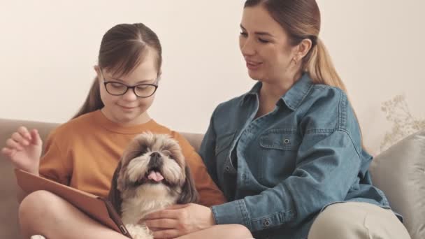 一个白种人的年轻女子 面颊上吻着她的小女儿 坐在沙发上与可爱的石子狗坐在一起 在数码平板电脑上看卡通片 — 图库视频影像