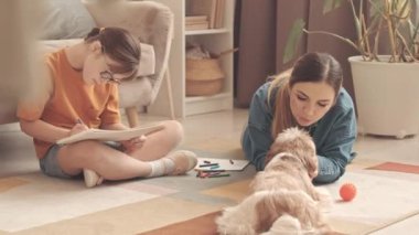 12 yaşında, beyaz bir kız. Oturma odasında halıya oturmuş resim çiziyor. Annesi de yakınlardaki sevimli köpekleriyle oynuyor.