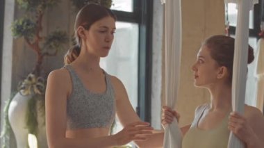 Parlak stüdyodaki ipek hamakta asılı dururken hava yogası antrenmanı sırasında antrenörle konuşan kızın orta yavaş hâli.