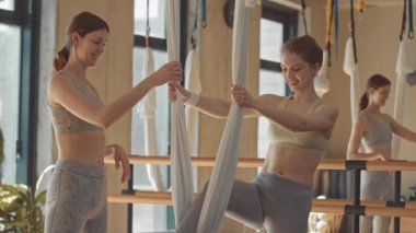 İpek hamakta öğrenciye yardım eden kadın eğitmen parlak geniş stüdyodaki hava yogası dersinde esneme egzersizi yapıyor.