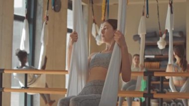 Yavaş yavaş hamakta oturan genç bayan antrenör grup yoga sınıfındaki kızlara kapalı alanda egzersiz gösteriyor.