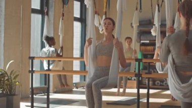 Gün ışığında geniş bir stüdyoda grup halinde hamaklarda oturan kızlara konuşan ve egzersiz yapan genç bayan eğitmen yavaşlığı.