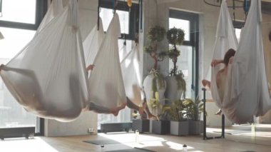 İpek hamaklarda havada asılı esneme egzersizleri yapan, geniş stüdyoda hava yogası yapan bir grup genç kadın.