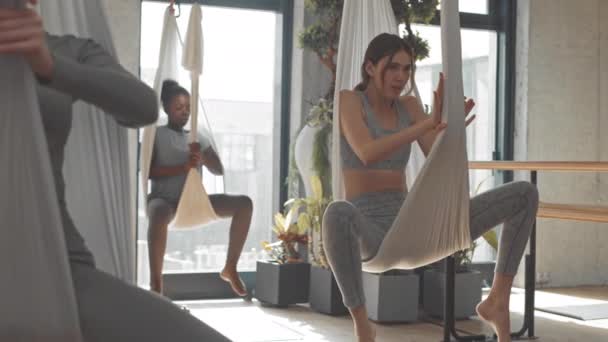 年轻美丽的空中瑜伽教练在丝绸吊床上向宽敞的演播室里的一群妇女展示运动 — 图库视频影像