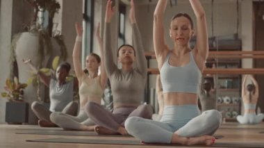 İçeride koçla yoga yapan, lotus pozu veren, dua eden elleri ve gözleri kapalı meditasyon yapan çok ırklı genç kadınların yavaşlaması.