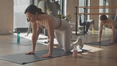 Kadın fitness eğitmeninin yavaşlığı ve çeşitli genç kadınlar gün ışığında stüdyoda kalçaları tekmeliyor.