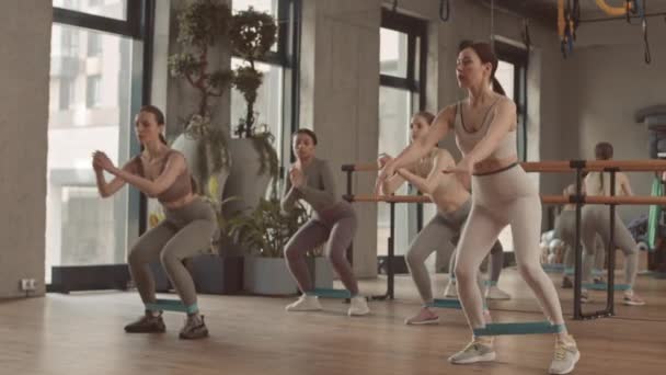 在明亮宽敞的演播室进行健身训练时 4名健康的年轻女子蹲在小腿肌肉上有阻力带的运动服的全长慢动作 — 图库视频影像