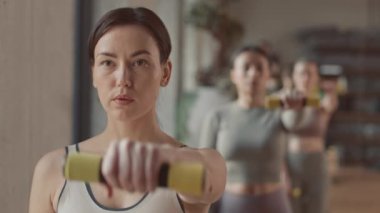 Dambıl gibi öne doğru yükselen genç bayan antrenör ve ardından tekrarlayan bir grup kadın spor salonunun içinde spor yapıyorlar.