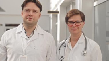 Modern hastane koridorunda kameraya gülümseyen beyaz erkek ve kadın doktor portresi.