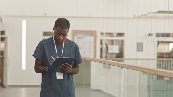 非洲裔美国男护士 身穿蓝色毛刷 手持剪贴板 对着站在医院走廊前的相机微笑的中等速度慢画像 — 图库视频影像