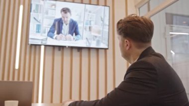 Yetişkin beyaz iş adamının yavaşlığı toplantı odasındaki duvarda asılı duran TV seti aracılığıyla çalışanlarıyla konuşuyor.