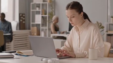 Çok ırklı meslektaşları arka planda bulanık bir şekilde etkileşime girerken, laptopta çalışan, odaklanmış, beyaz bir kadının orta yavaşlığı.