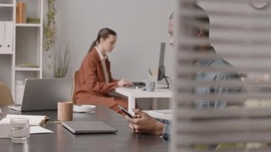 İş yerindeki iş yerinde iş arkadaşı bilgisayarla uğraşırken, kot tişörtlü, akıllı telefonuyla mesajlaşan genç siyahi bir adamın orta yavaşlığı.
