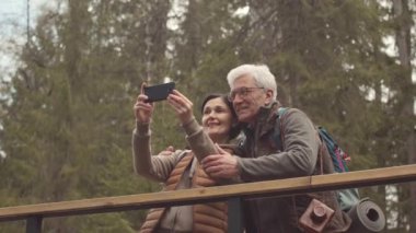 Ormanda yürüyüş yaparken akıllı telefondan selfie çeken olgun, aktif bir çiftin yavaşlaması.