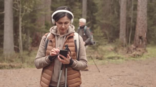 中等速度的成熟白人女人 站在森林里的小径上 头戴耳机 在智能手机上打滚 老年人北欧人走来走去 — 图库视频影像