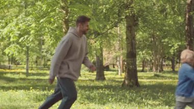 PAN yavaş Kafkasyalı adam ve 7 yaşındaki oğlu güneşli bir günde parkta eğleniyor, ağaçların etrafında kovalamaca oynuyorlar.