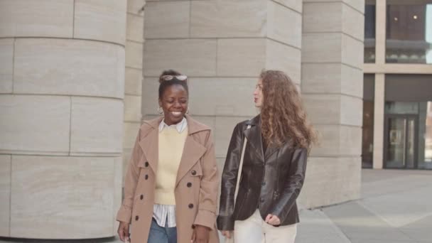 市内中心部を歩く2人の若い魅力的な多様な女性の中低速 近代的なオフィスビルを見て会話を持つ — ストック動画