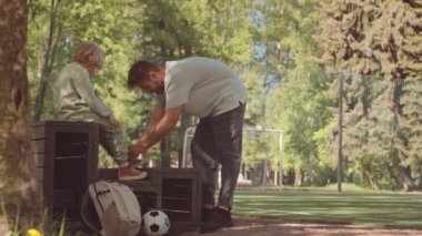 Güneşli bir günde parkta birlikte futbol oynamadan önce oğullarının ayakkabılarını bağlayan bir babanın yavaşlığı.