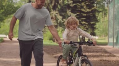 Parkta bisiklet süren neşeli küçük bir çocuk ve onu destekleyen şefkatli babası dengesini korumasına yardım ediyor.