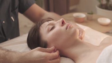 Spa salonundaki kanepeye uzanmış güzel genç bir kadının kulak memelerine masaj yapan tanınmayan masörün yavaş yavaş yakın çekimi.