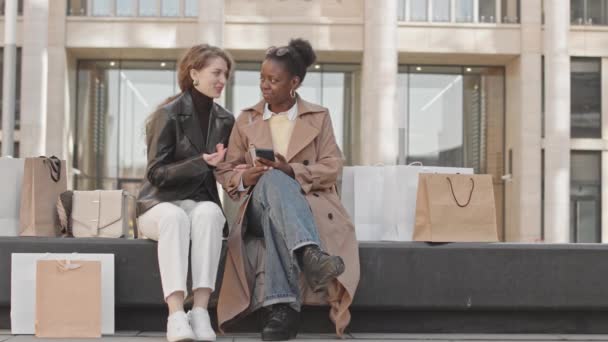2人の若いスタイリッシュな多民族のガールフレンドのスローモは スマートフォンを見て 買い物袋の多くと屋外のベンチに座っている間にチャット — ストック動画