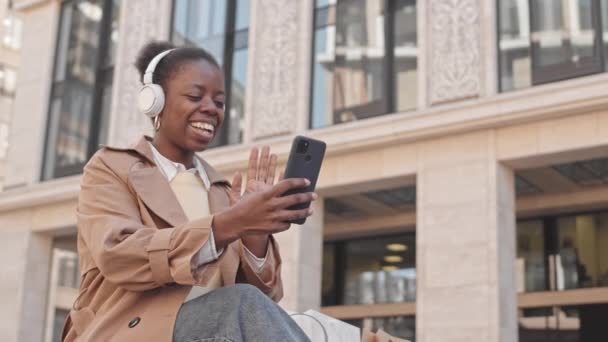 一个快乐的年轻黑人女人坐在户外长椅上 坐在智能手机上与朋友聊天 用耳机视频显示她在购物袋里购物的情景 — 图库视频影像
