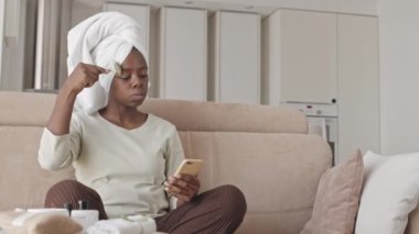 Kafasında havluyla akıllı telefondan kayarken, evinde koltuğunda otururken, kuvars silindirli yüz masajı yapan güzel Afro-Amerikan kadının orta yavaş görüntüsü.