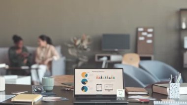 Pazarlama istatistikleri olan dizüstü bilgisayar, smm uzmanının ofis masasında ekranda. Bulanık arka planda sohbet eden iki farklı bayan arkadaş.