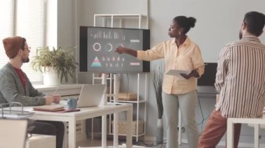 Afrika kökenli Amerikalı kadın pazarlama uzmanı, iş grafikleri ve çizelgeleri ekranda gösterirken aynı zamanda meslektaşlarına ürün sunumu yapıyor.