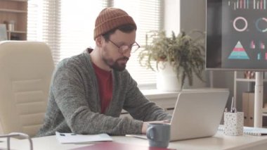 Ofis masasında laptopta çalışan günlük giysili ve bereli beyaz bir adam.