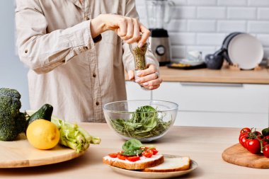 Sebzeli salataya baharat katan genç bir kadın. Mutfakta masada duruyor ve akşam yemeği hazırlıyor.