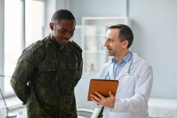 stock image Mature Caucasian doctor in white coat showing good checkup results to young Black military officer pn digital tablet screen