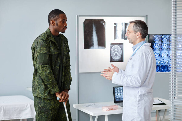 Young Black military man leaning on walking stick discussing something with mature Caucasian doctor after doing x-ray